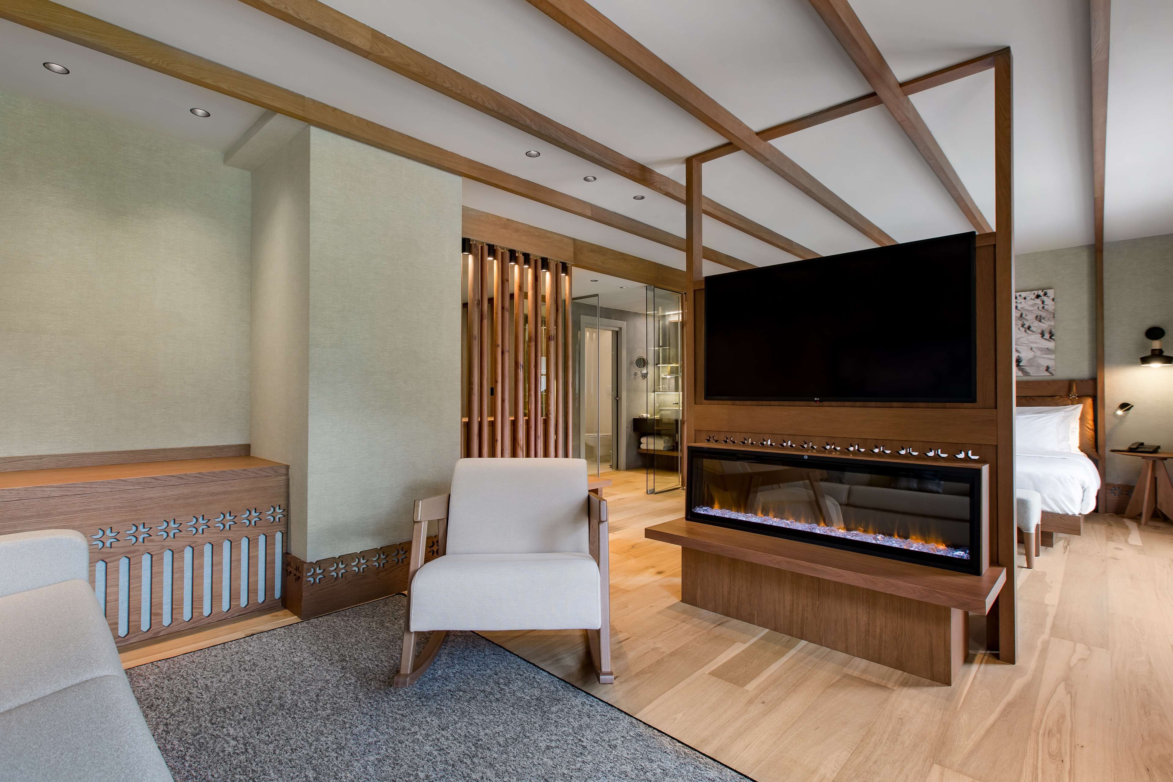 A modern hotel room with a large flat-screen TV, a fireplace, a comfortable chair, and a wooden ceiling structure.