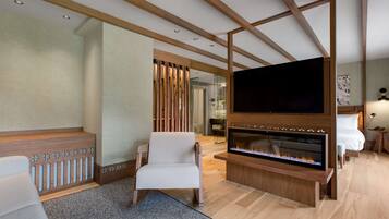 A modern hotel room with a large flat-screen TV, a fireplace, a comfortable chair, and a wooden ceiling structure.