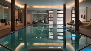 En modern inomhuspool med väggar av genomskinligt glas, träkolonner och sittområden.