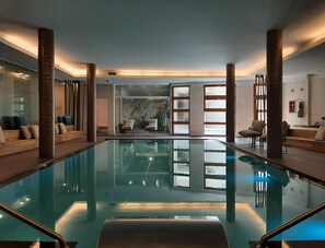 Une piscine intérieure moderne avec des parois en verre transparent, des colonnes en bois et des espaces aménagés pour se détendre.