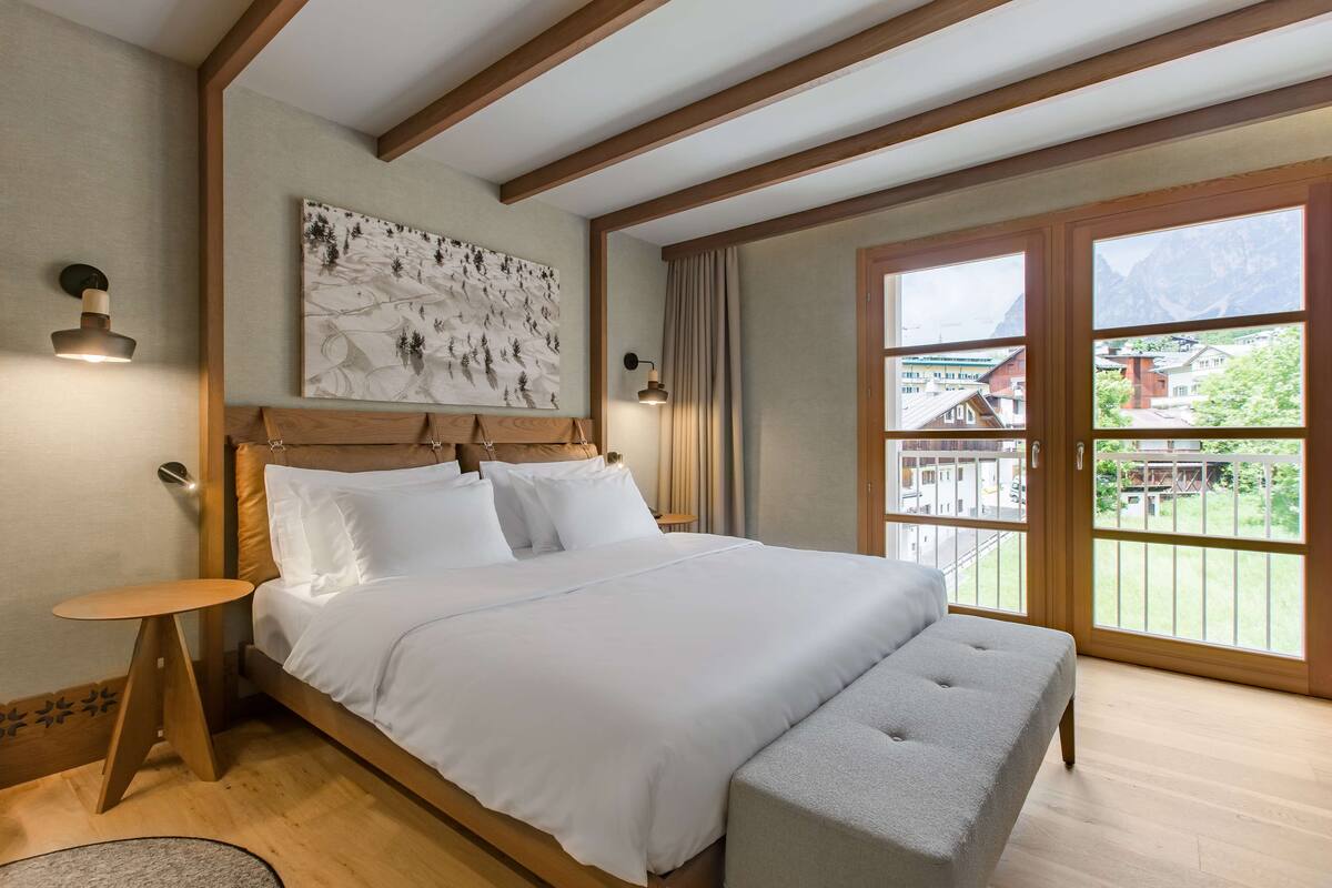 Una camera da letto moderna con una testiera in legno, un letto grande con lenzuola bianche, un comodino e una panca. Una grande finestra offre vista su edifici e montagne.