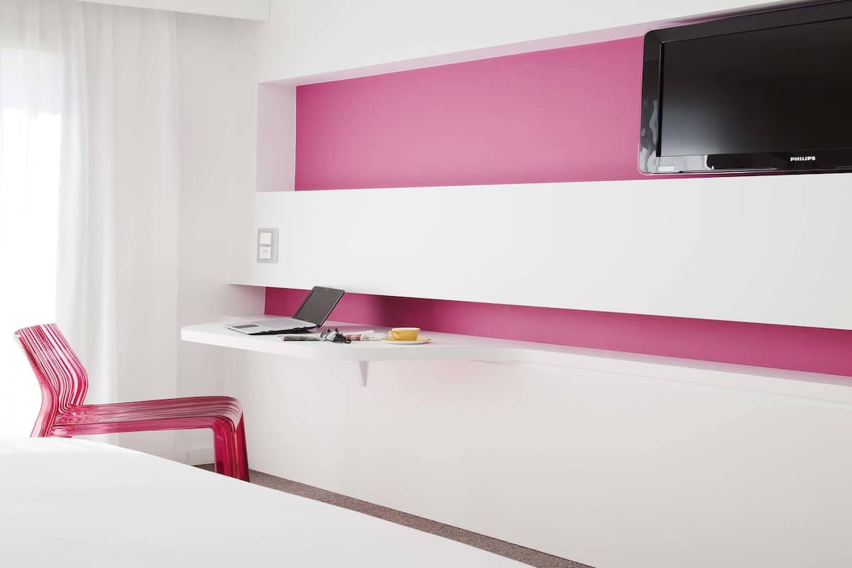 A modern hotel room with a white desk, a laptop, a flat-screen TV, and a red chair.