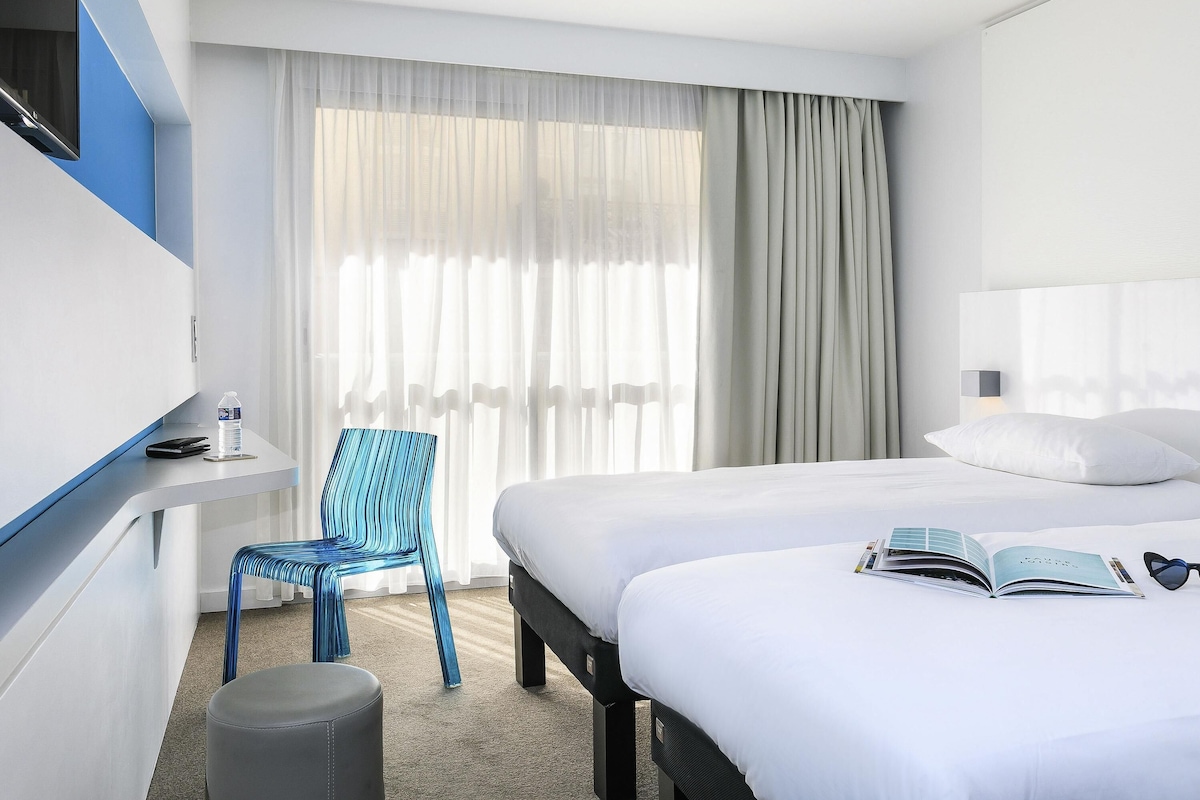 Una habitación de hotel con una cama, una silla, una mesita con una botella y un libro sobre la cama.