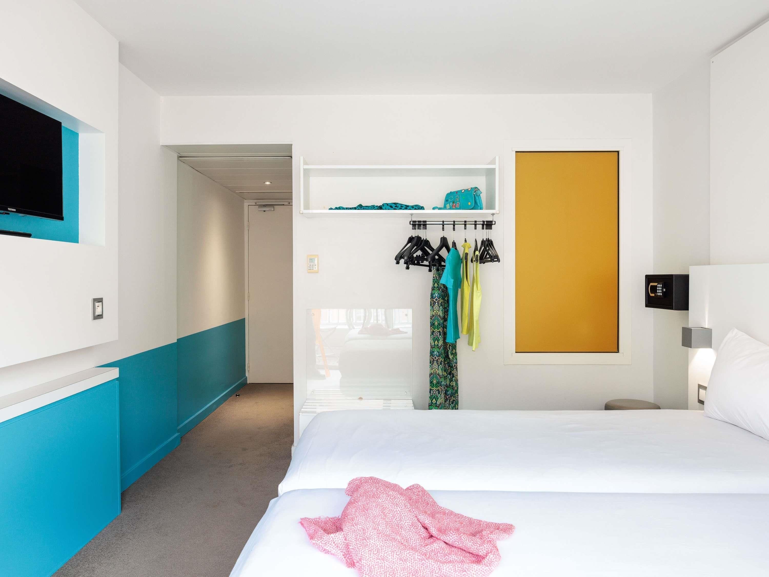Una habitación de hotel con cama, televisor en la pared, perchero con ropa y armario.