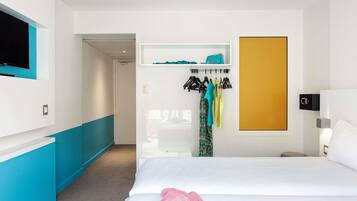 Una habitación de hotel con cama, televisor en la pared, perchero con ropa y armario.