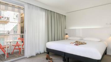 A hotel room with a bed, a nightstand, a lamp, a balcony with chairs, and a view of buildings.