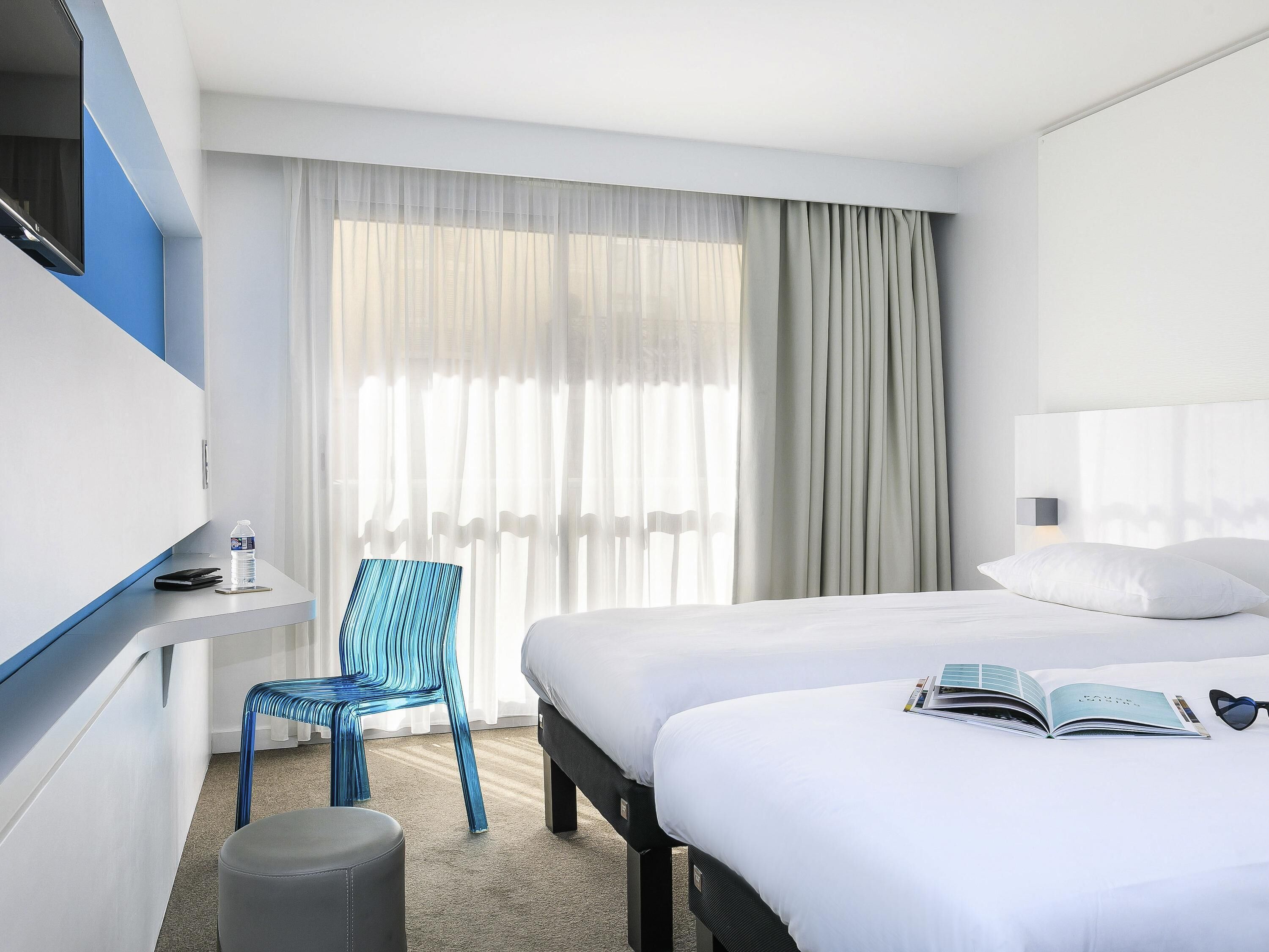 Una habitación de hotel con una cama, una silla, una mesita con una botella y un libro sobre la cama.