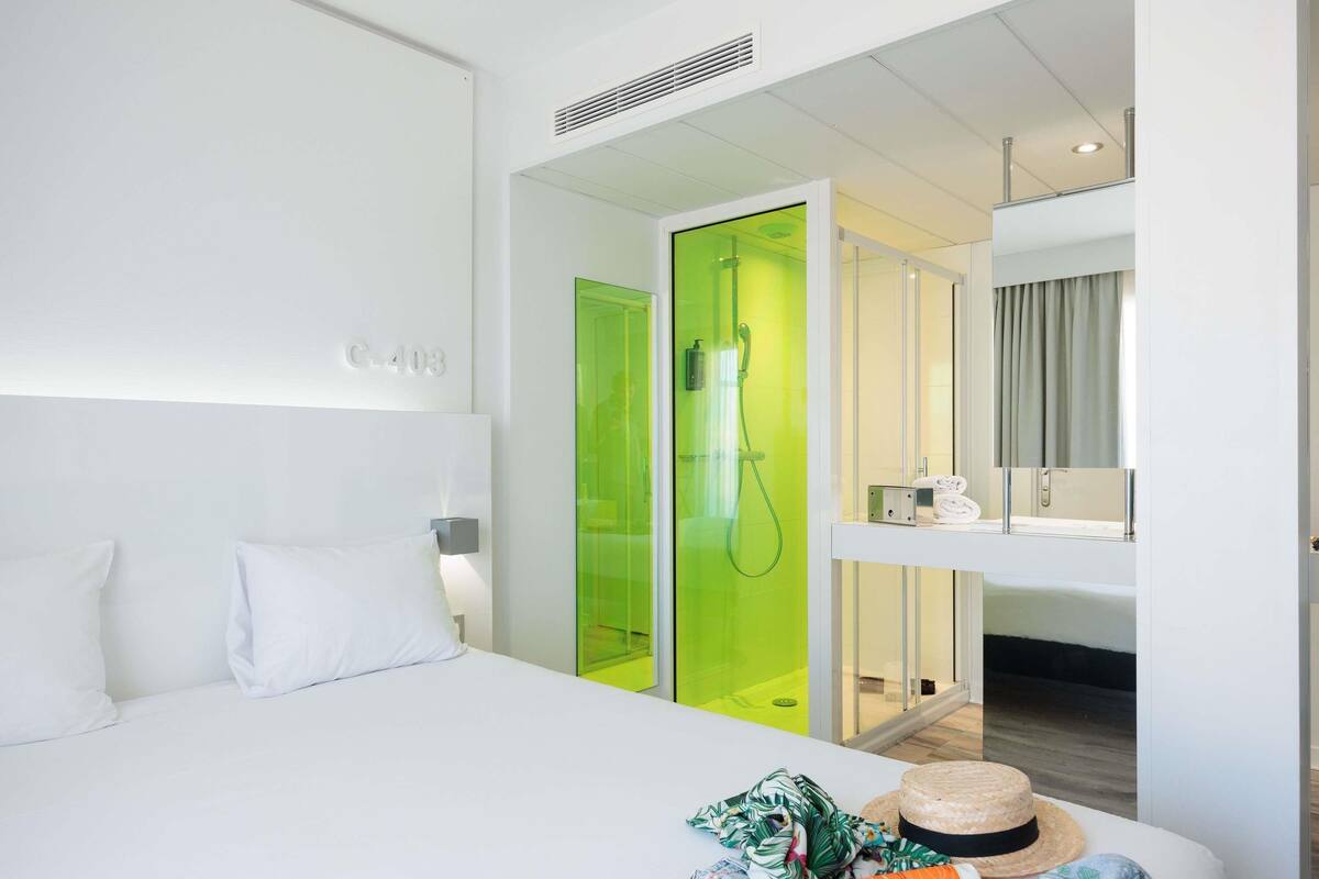 A modern hotel room with a bed, a green shower enclosure, a bathroom with a mirror, and a window with curtains.