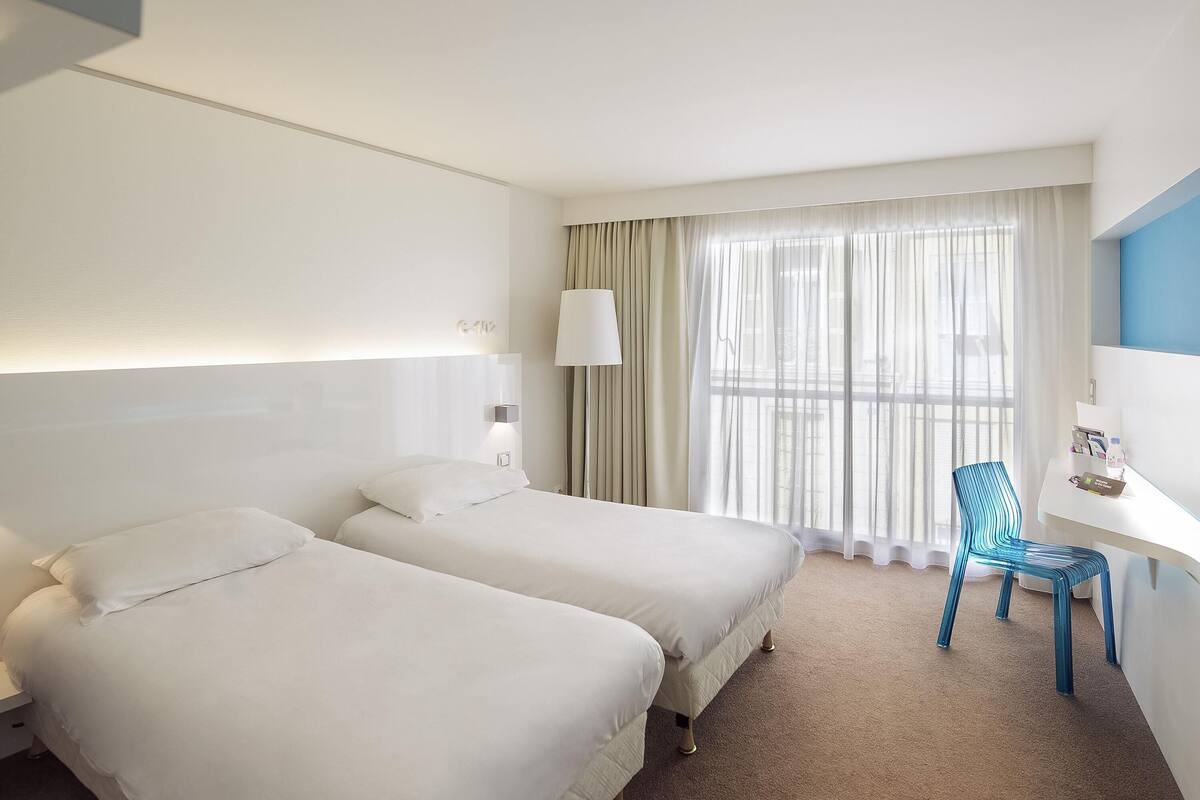 A hotel room with two beds, a desk, a chair, a lamp, and a window with curtains.
