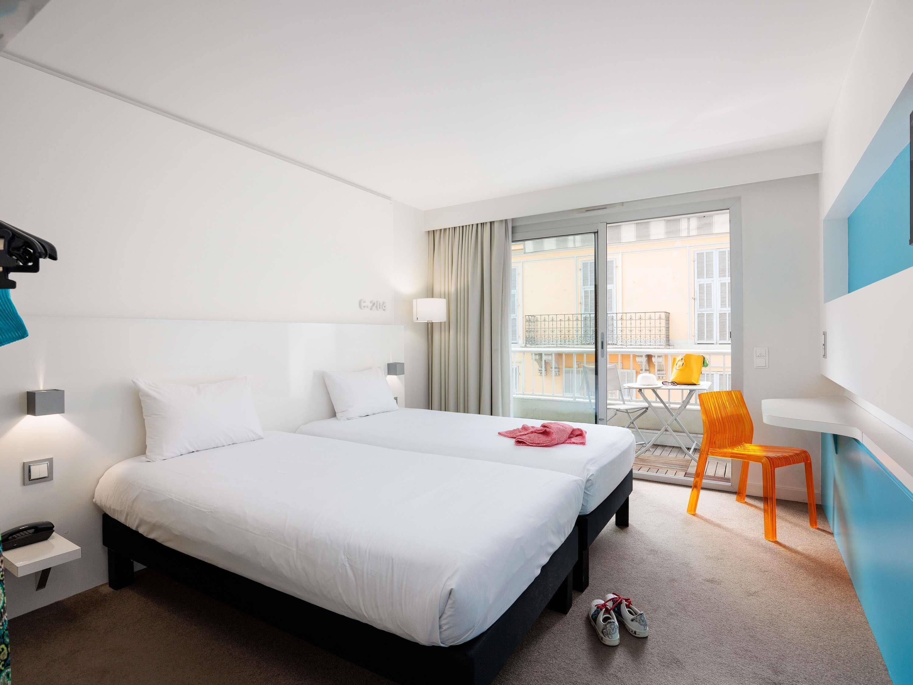 Een hotelkamer met een tweepersoonsbed, een klein tafeltje met een stoel, een balkon met uitzicht op gebouwen en een schoenenrek.