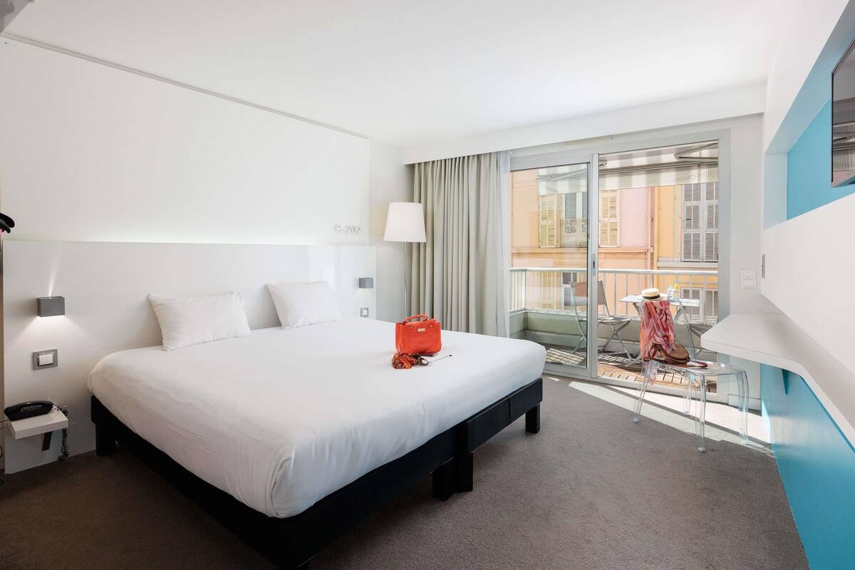 A hotel room with a large bed, a desk with a telephone, a lamp, and a view of the outside balcony.