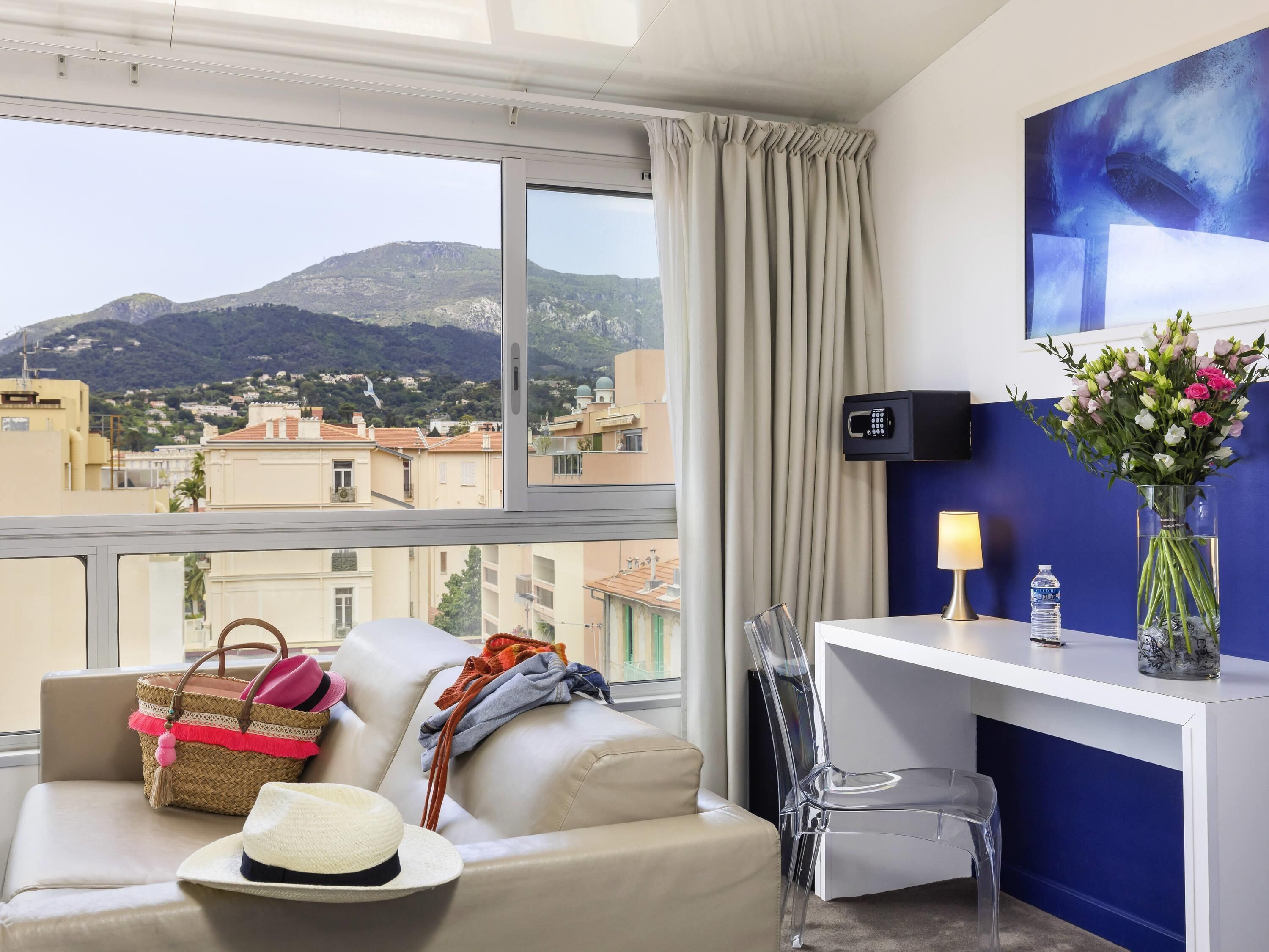 Habitación de hotel con vistas a montañas, un sofá, una silla transparente, un escritorio con lámpara, un jarrón con flores y una botella de agua.