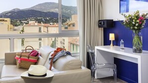 A hotel room with a sofa, a desk, a chair, a vase with flowers, a lamp, a water bottle, and a view of a mountain and buildings. - ibis Styles Menton Centre (Menton)