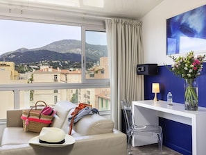Habitación de hotel con sofá, escritorio, silla, jarra con flores, lámpara, botella de agua y vistas a una montaña y edificios.