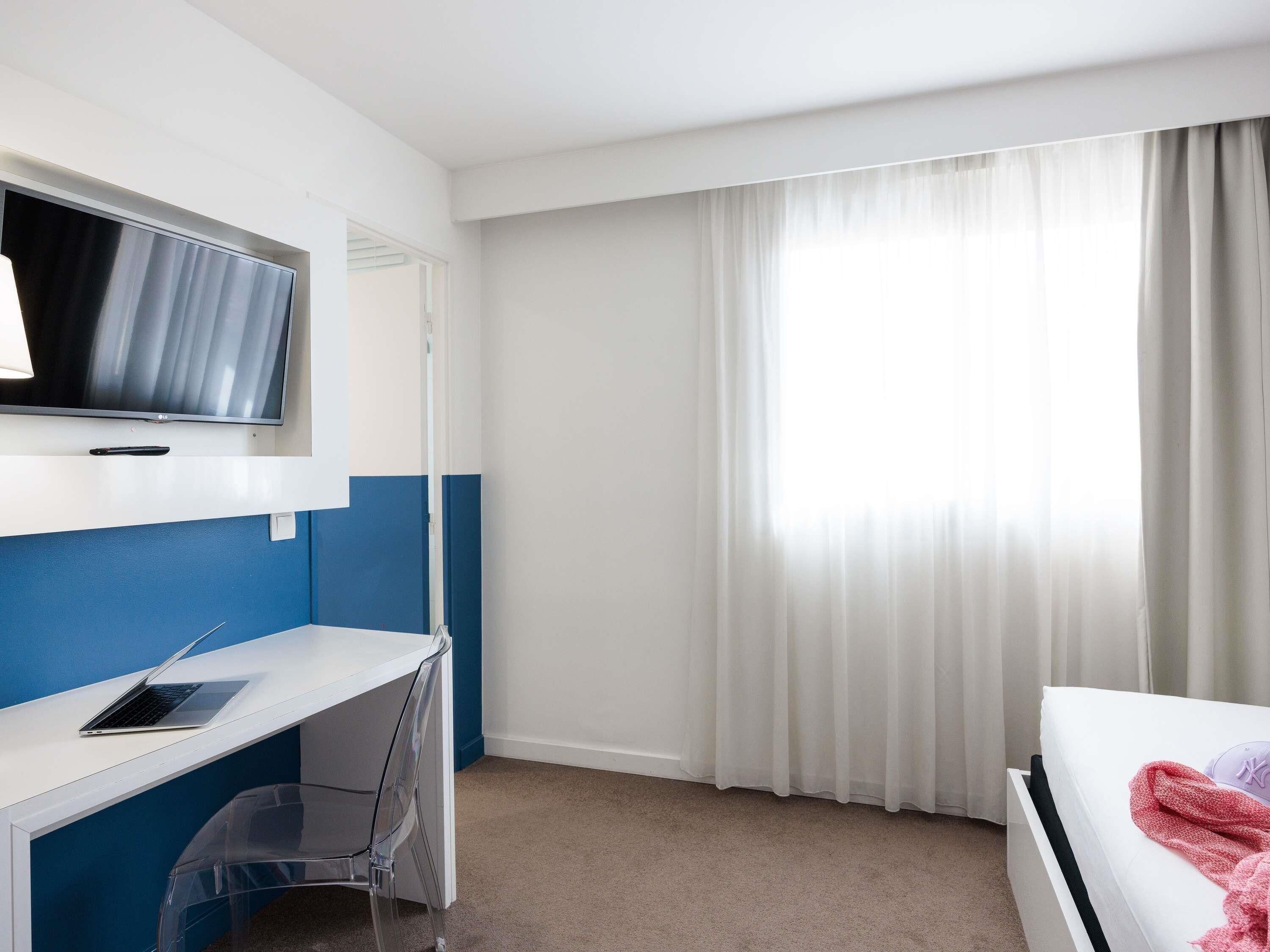 A hotel room with a bed, a desk with a laptop, a TV, and a window with curtains.