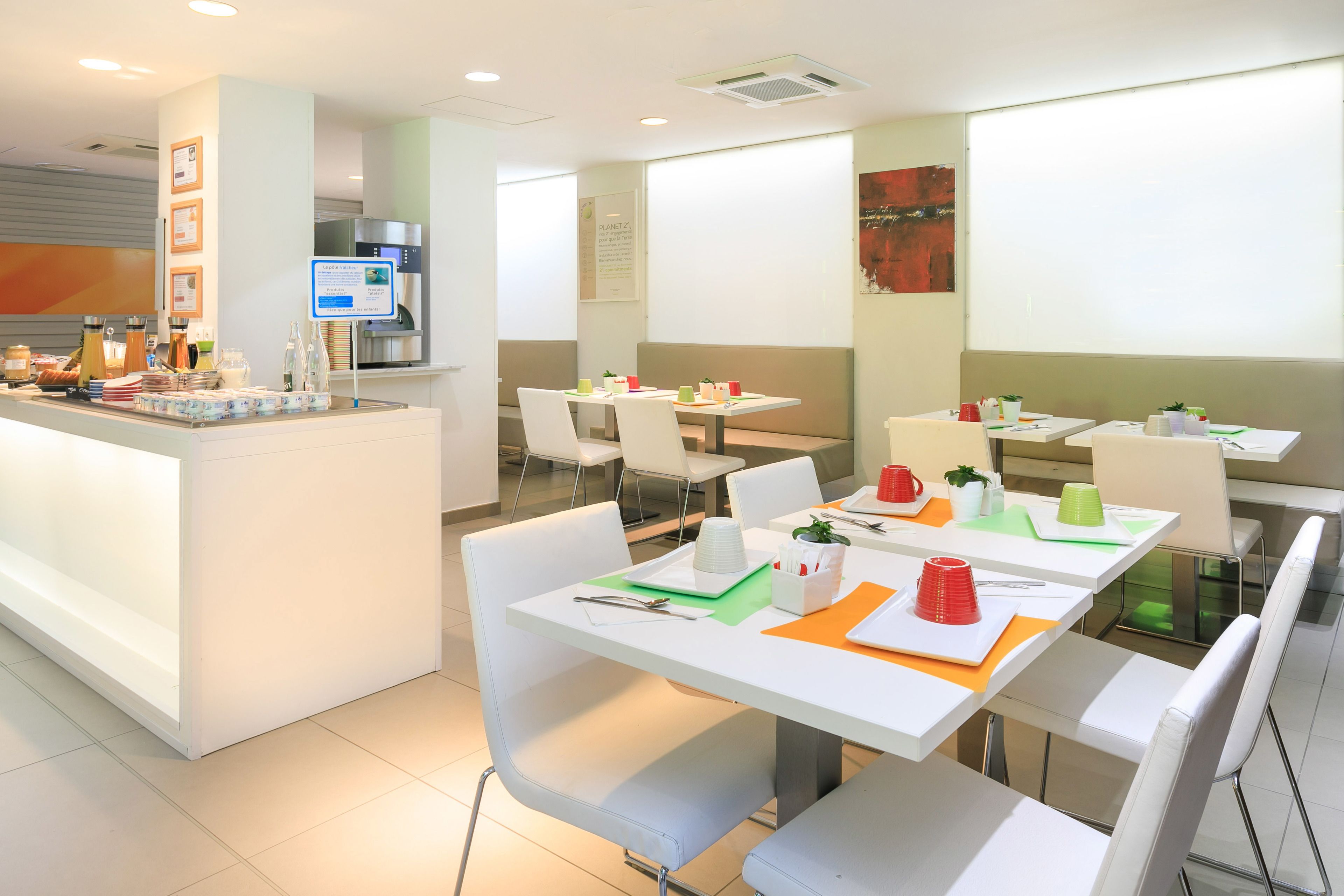 Un comedor moderno con mesas y sillas blancas, una barra de buffet con comida y bebidas, y una pantalla de proyección en la pared.