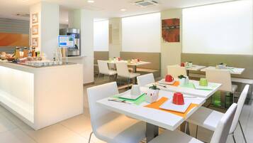 Un comedor moderno con mesas y sillas blancas, una barra de buffet con comida y bebidas, y una pantalla de proyección en la pared.