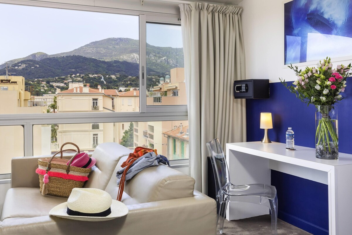 Ein Hotelzimmer mit Blick auf Berge, einer Couch, einem transparenten Sessel, einem Schreibtisch mit Lampe, einer Vase mit Blumen und einer Wasserflasche.