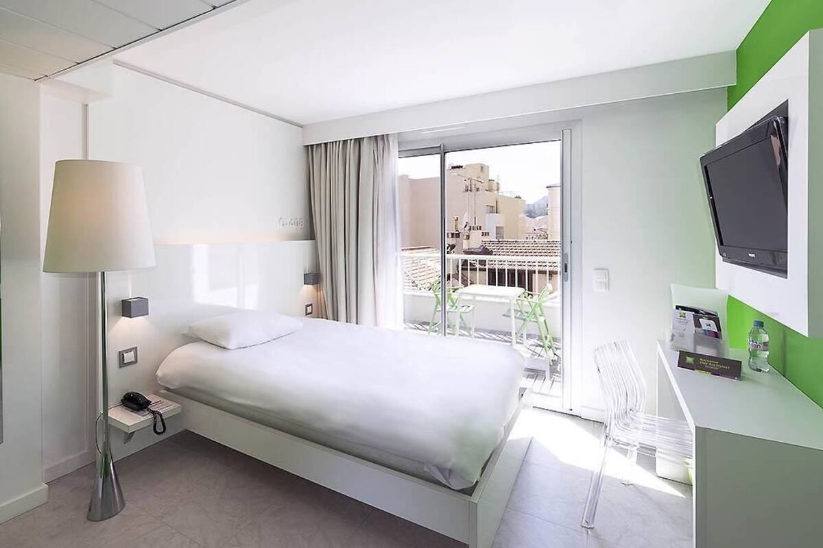 A modern hotel room with a bed, bedside lamp, and a view of buildings through a large window.