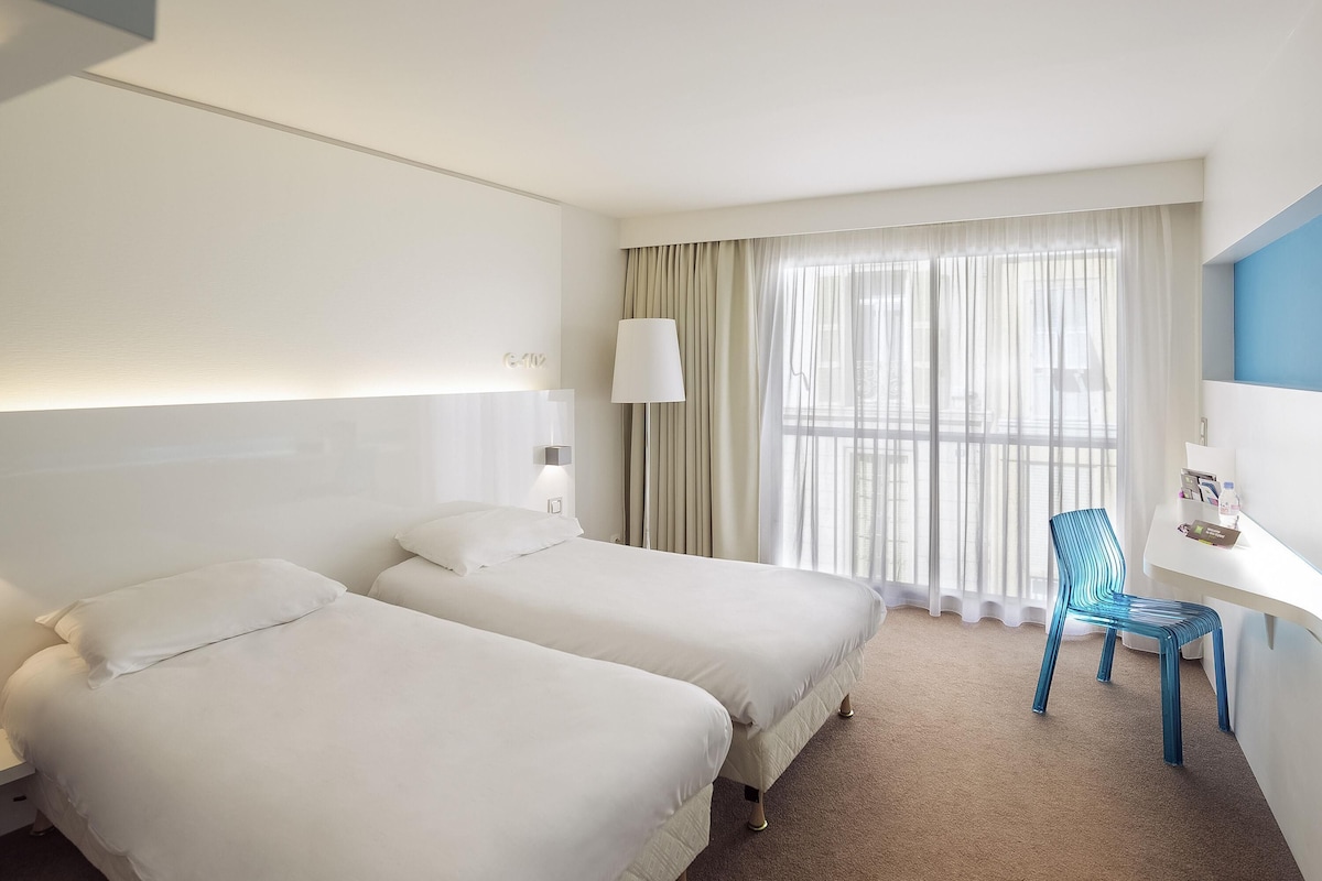Habitación de hotel con dos camas, un escritorio, una silla, una lámpara y un ventanal con cortinas.