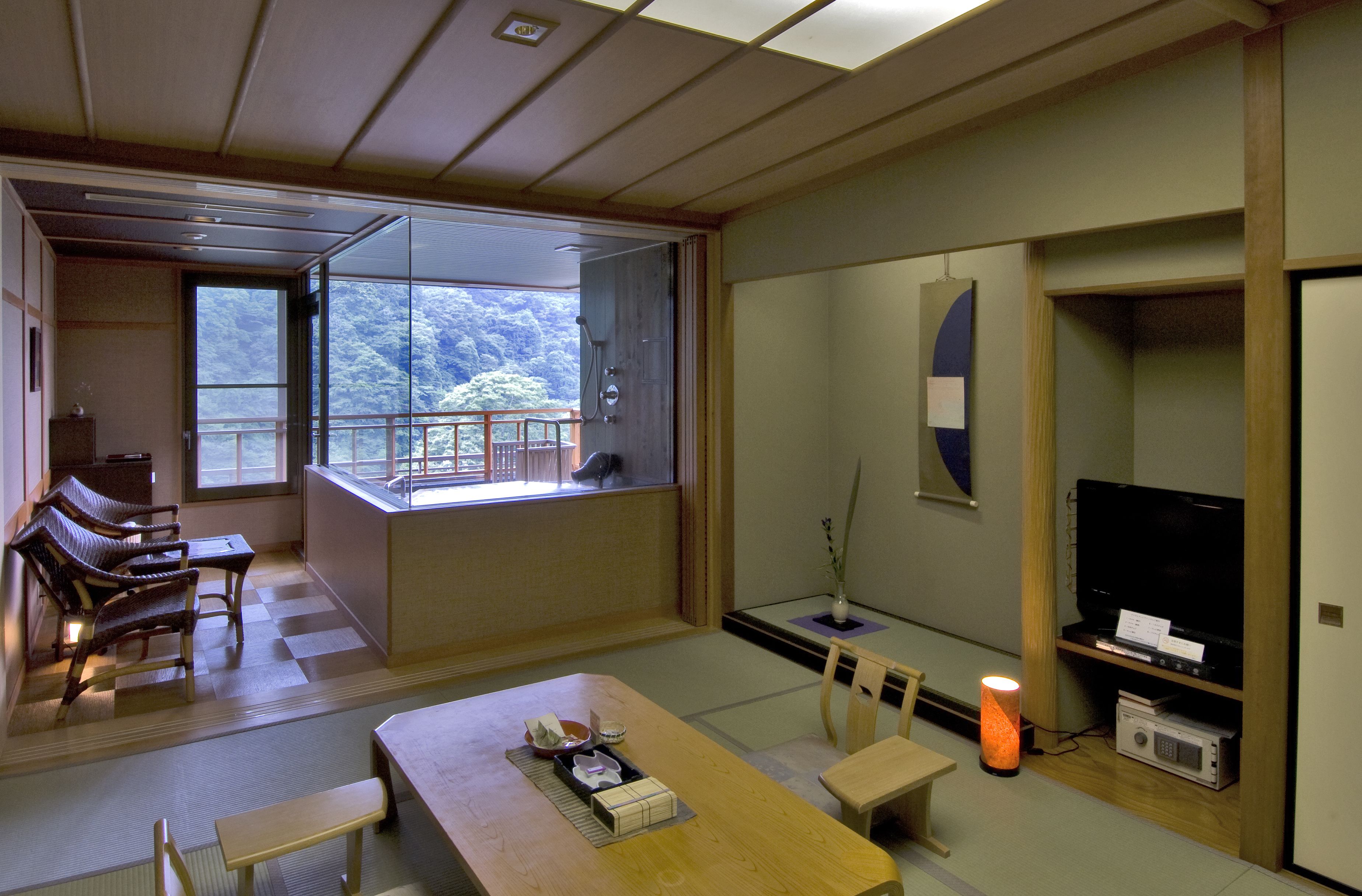 Een traditionele Japanse kamer met tatami vloerbedekking, een lage tafel met kussens, een televisietoestel en een schuifdeur die leidt naar een balkon met uitzicht op bomen.