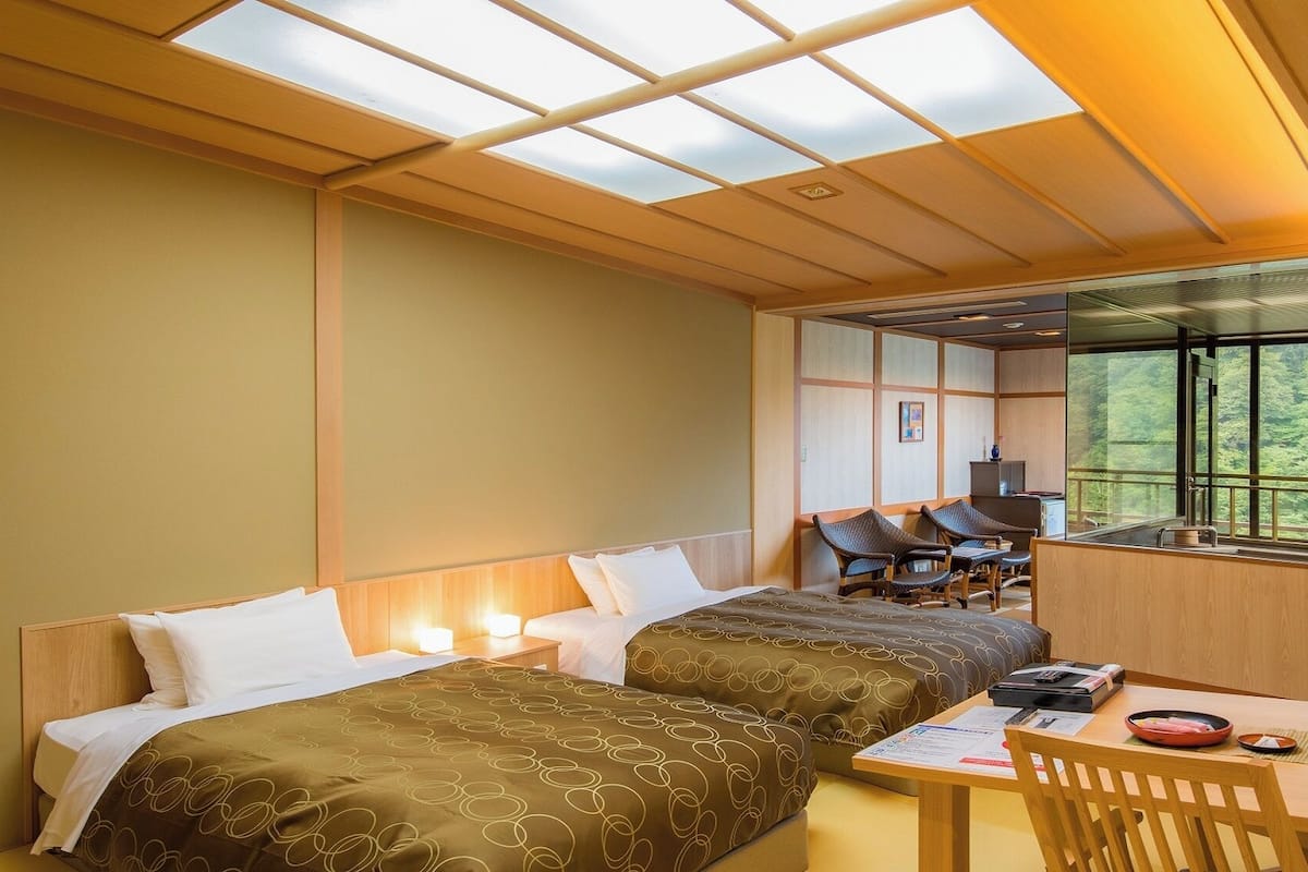 A hotel room with two beds, a dining table, and a view of a forest through a large window.
