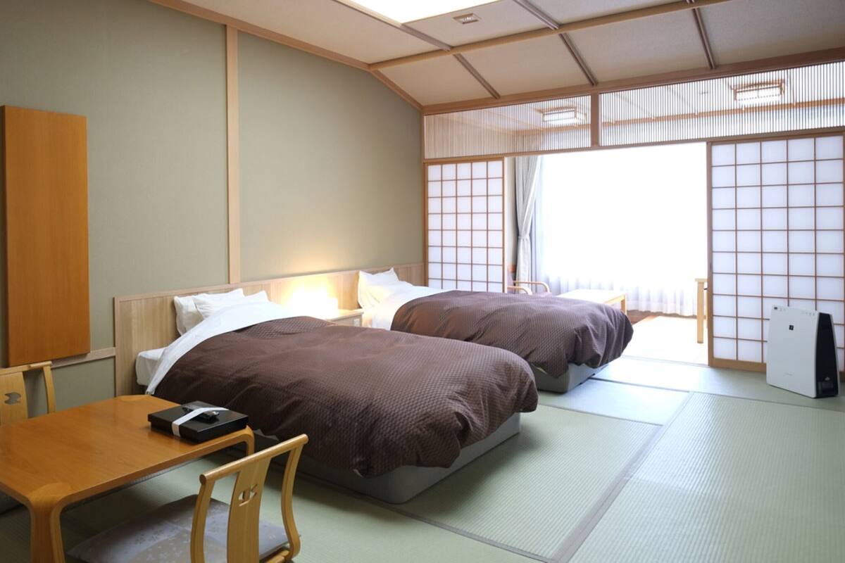 A traditional Japanese hotel room with tatami flooring, two beds with brown bedding, a wooden desk, and a shoji screen door.