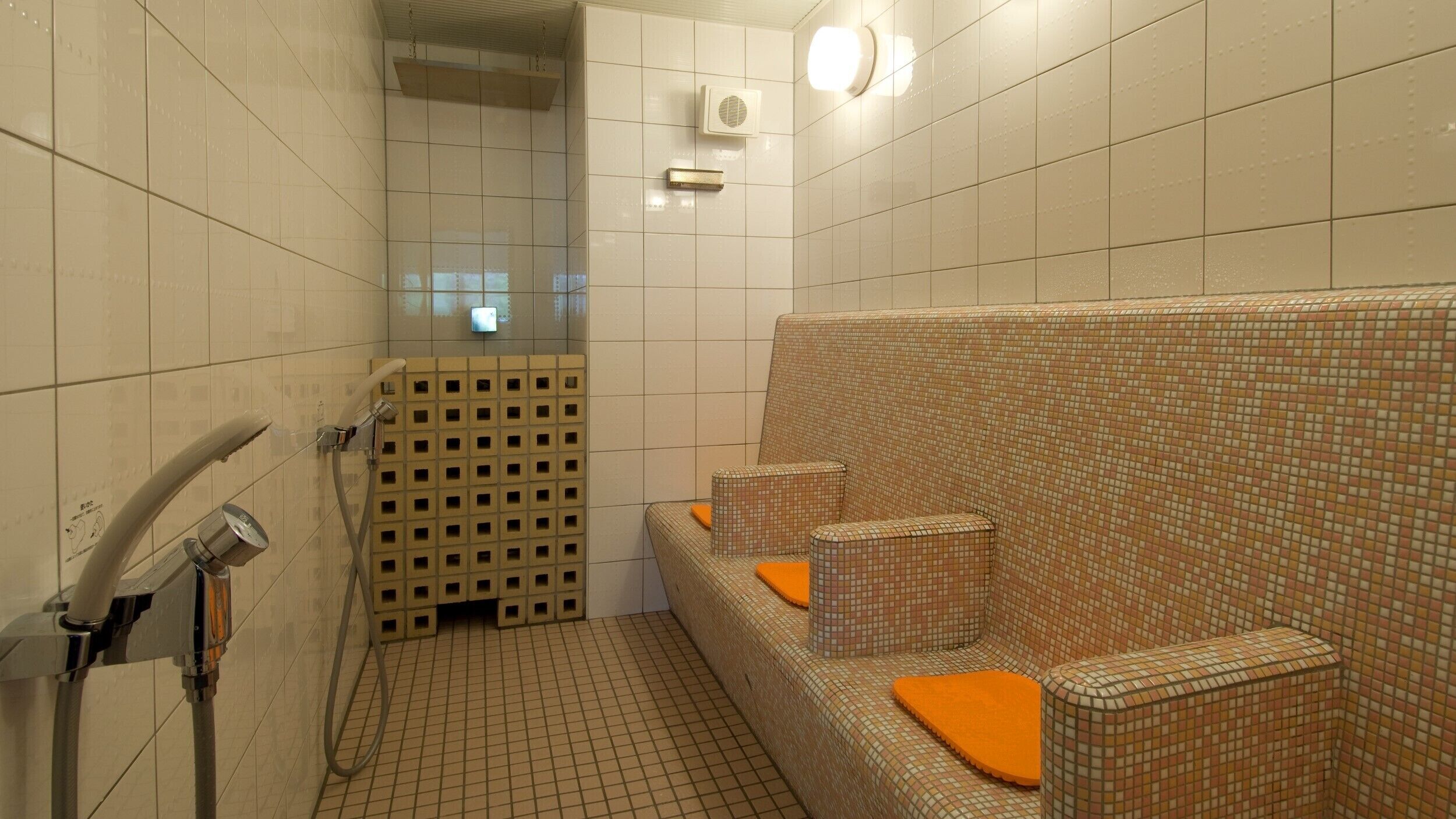 A bathroom with tiled walls, a shower area, and a seating area with orange cushions.