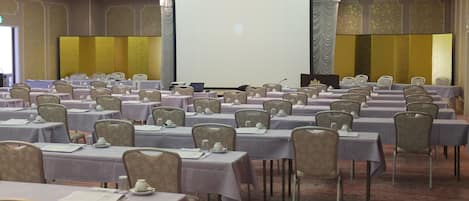 Un salón de conferencias con filas de mesas, sillas y una pantalla grande.