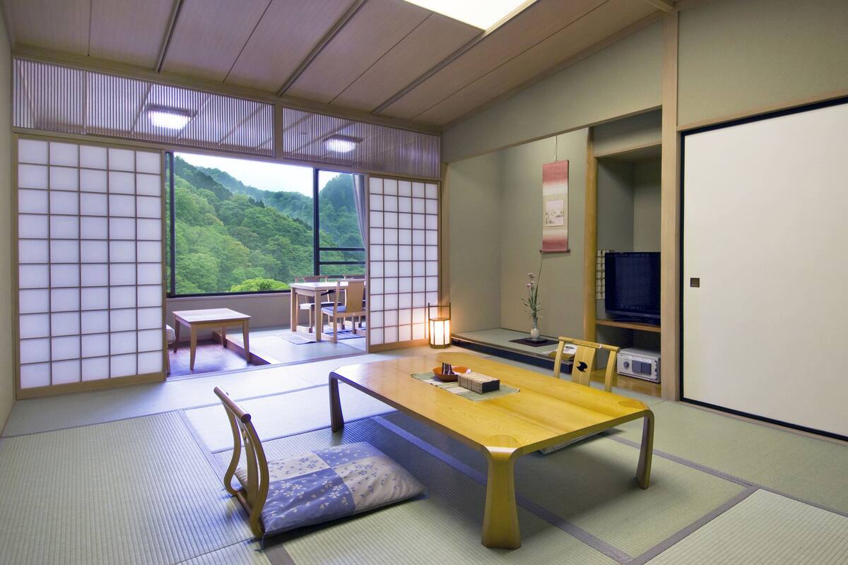 A traditional Japanese room with tatami flooring, a low table, chairs, a television, and a view of a lush green landscape.