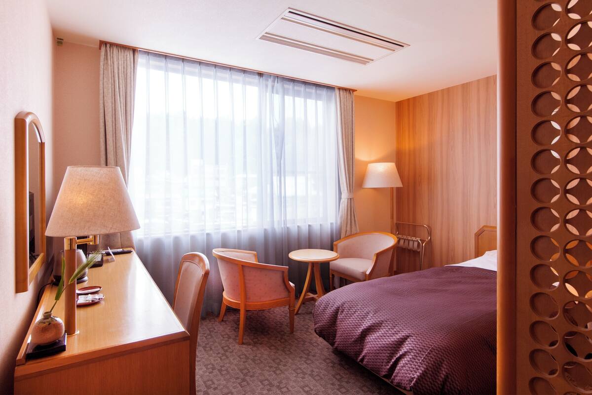 A hotel room with a bed, two chairs, a desk with a lamp, a bedside table, and a large window with curtains.