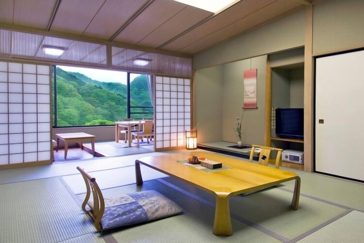 A traditional Japanese room with tatami flooring, a low table, a TV, and a view of a green landscape.