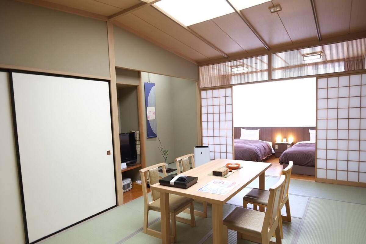 A traditional Japanese-style room with tatami flooring, a low table with chairs, a bed with a purple bedspread, and a sliding door.