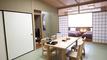 Ein traditioneller japanischer Raum mit Tatami-Böden, einem niedrigen Tisch mit Stühlen, einem Bett mit violetter Bettdecke und einer Schiebetür.