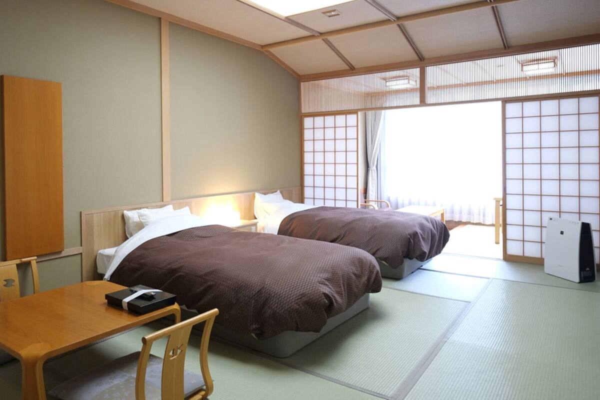 A traditional Japanese room with tatami flooring, two beds with brown bedding, a wooden table, and a small desk with a computer.