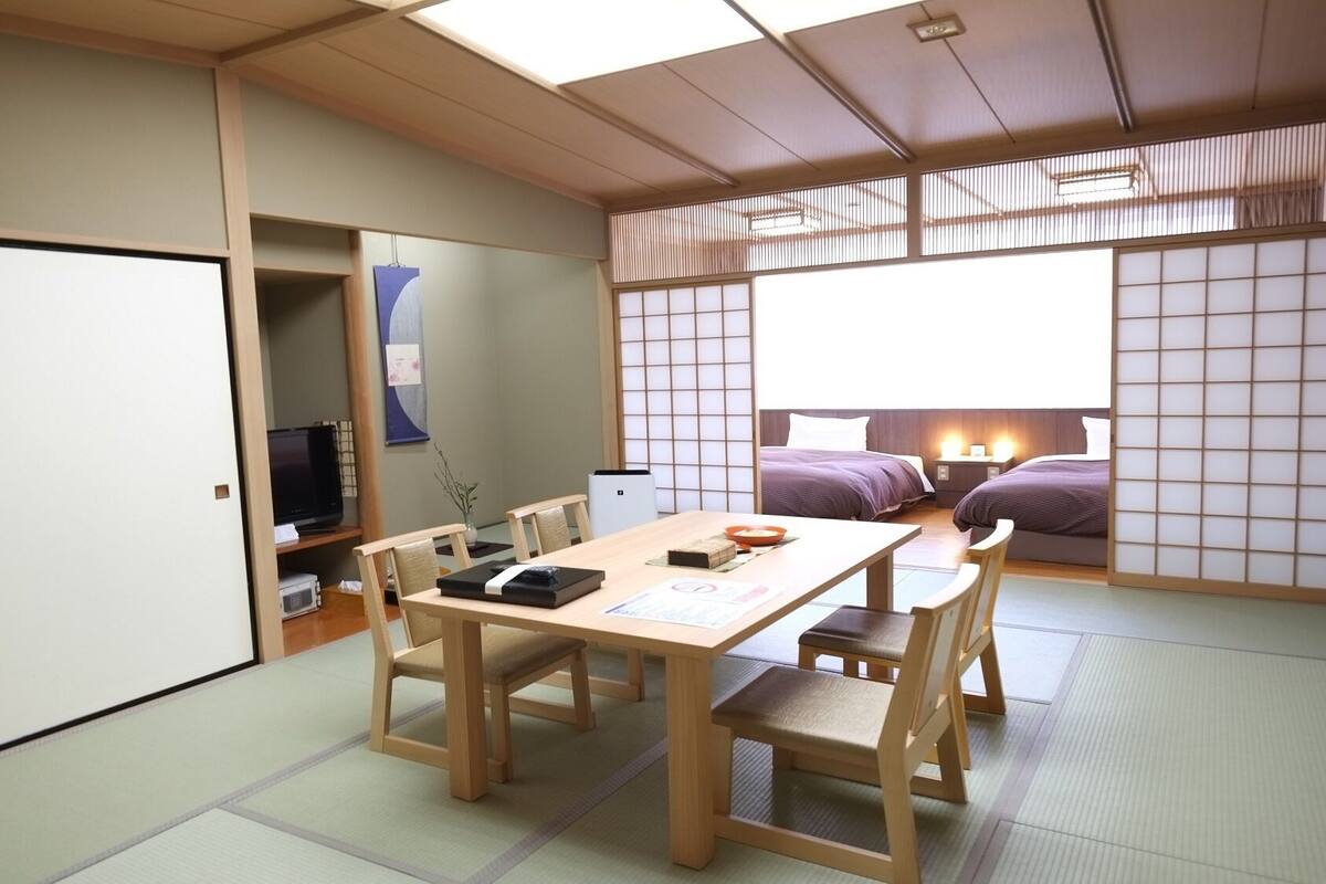 A traditional Japanese-style room with tatami flooring, a low table with chairs, a bed with purple bedding, and sliding doors.