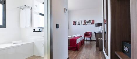A hotel room with a large window, a bed with a red bedspread, a chair, a desk with a lamp, and a wall-mounted artwork.