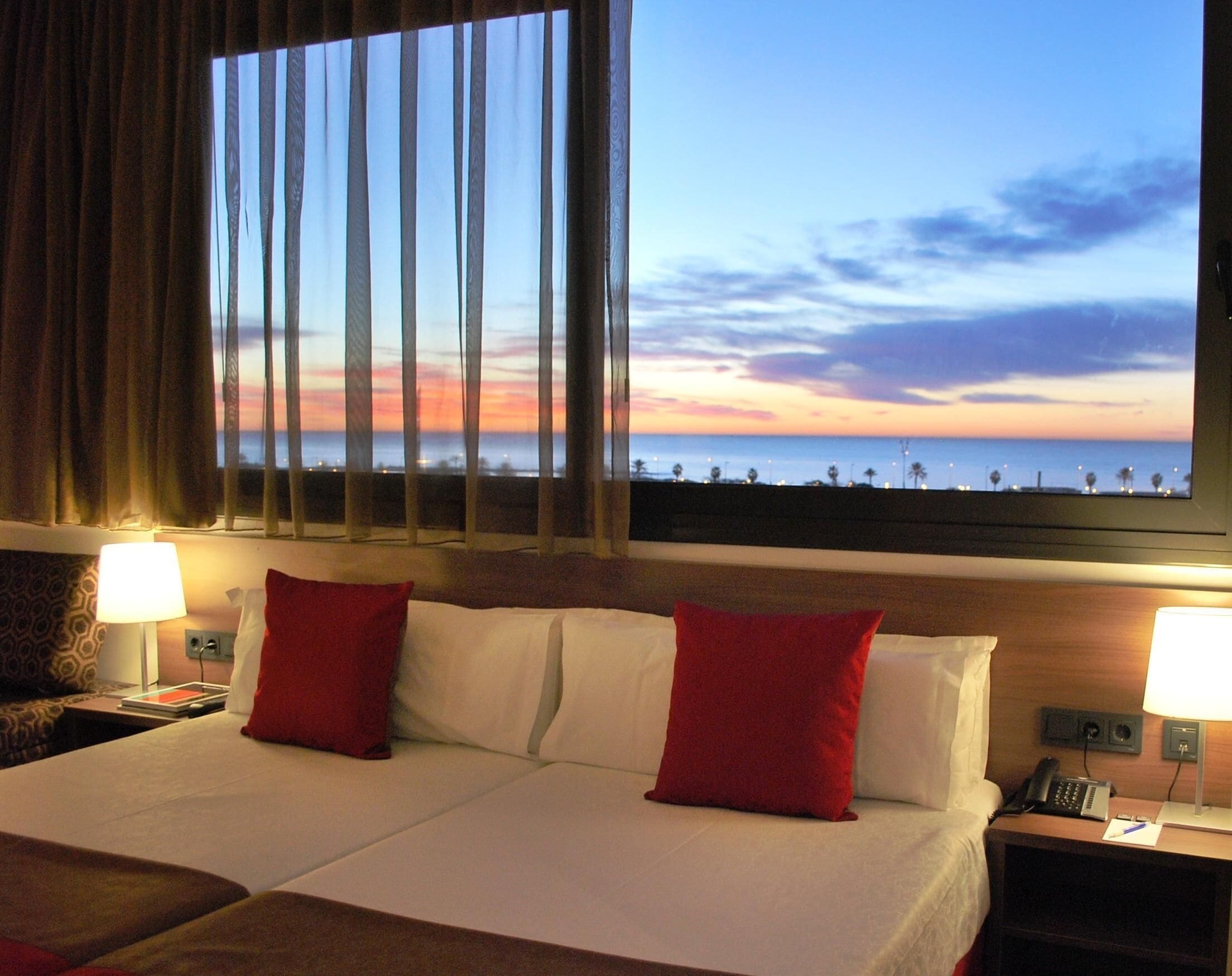 A hotel room with a bed, two red pillows, a nightstand with a phone, and a view of the beach at sunset.