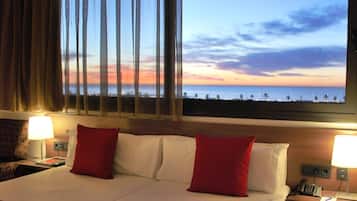 Habitación de hotel con una cama, dos almohadas rojas, una mesita de noche con un teléfono y vistas a la playa al atardecer.