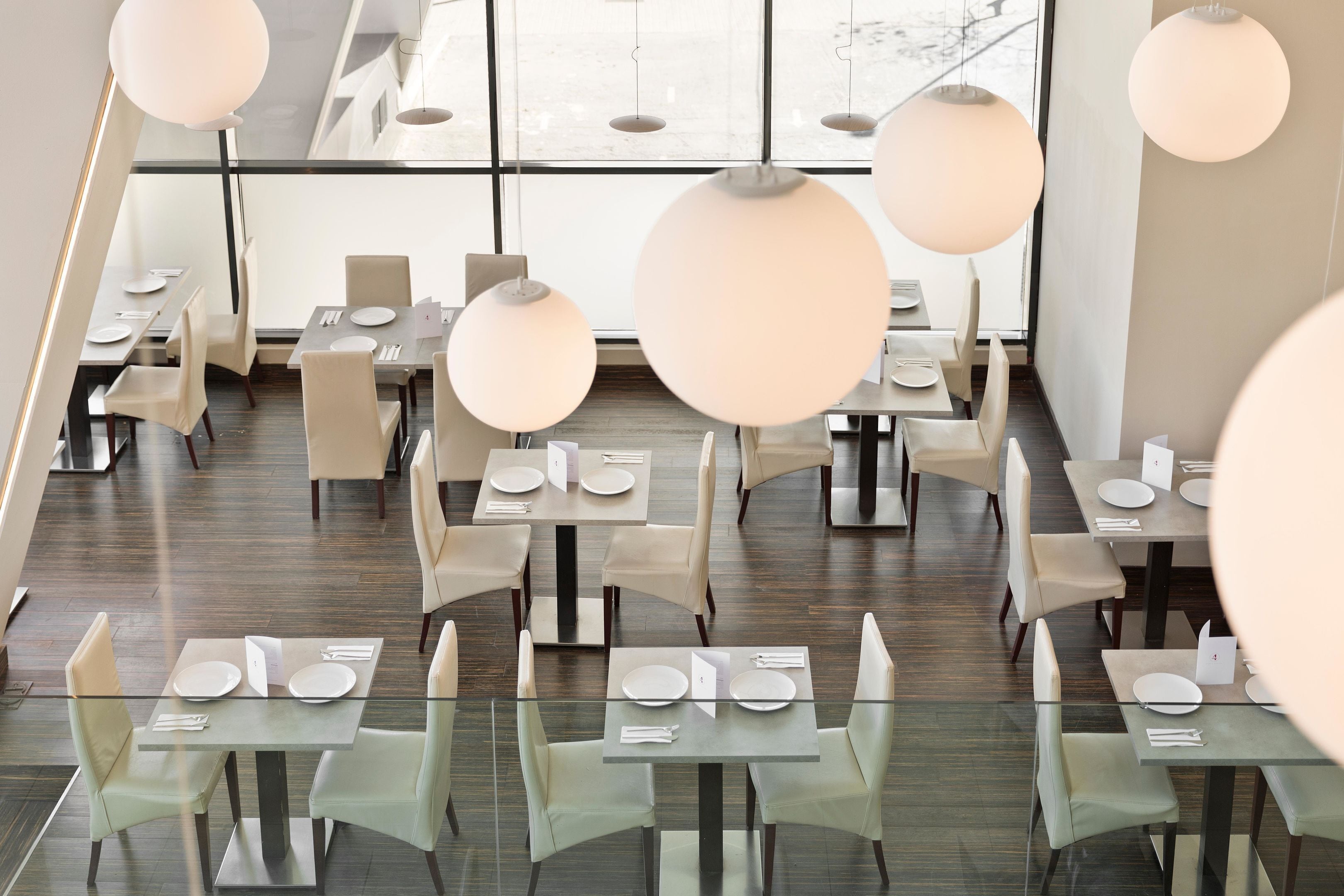 A modern restaurant with glass tables and chairs, large pendant lights, and a view through large windows.