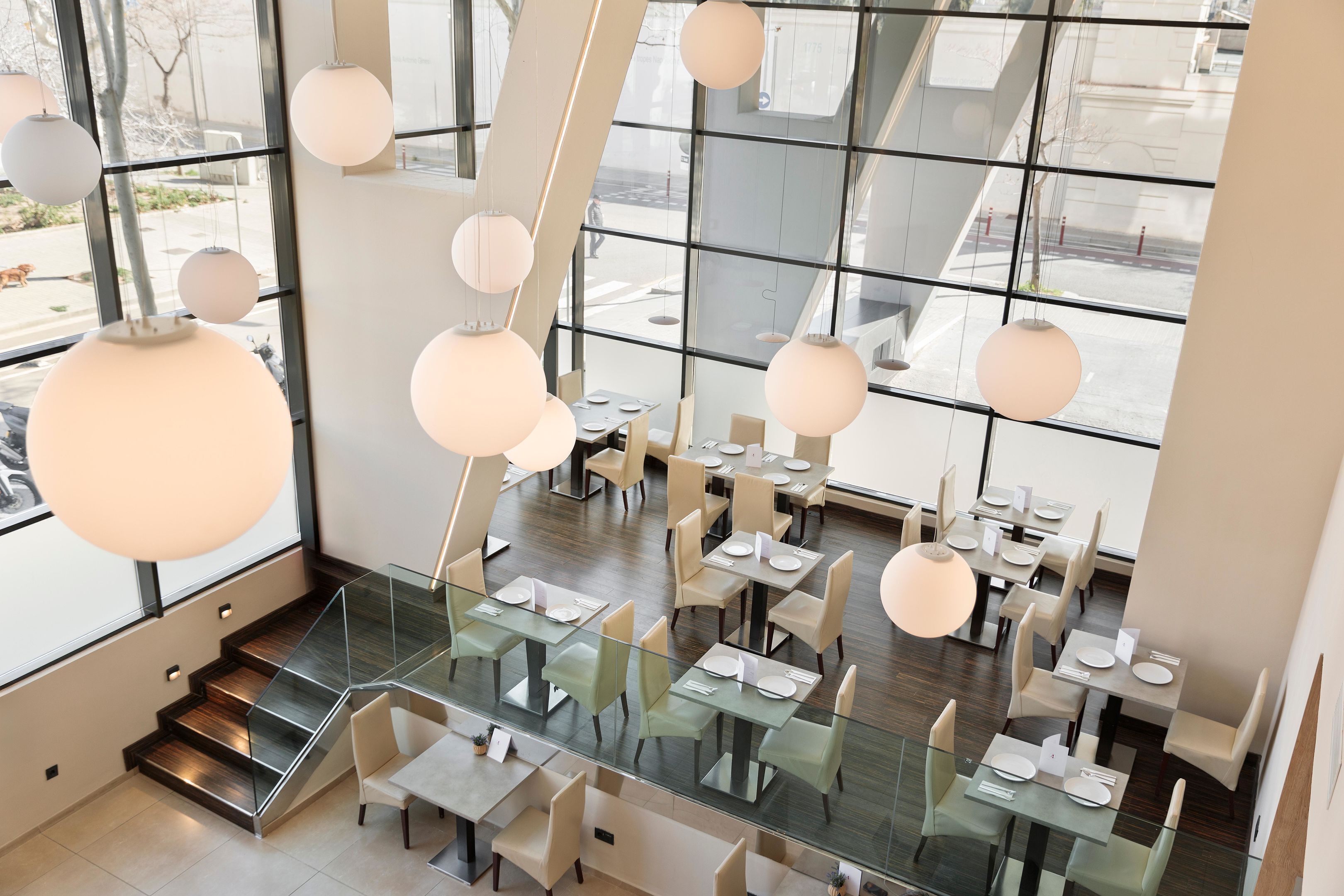 Un restaurante moderno con mesas y sillas de vidrio, ventanas amplias y lámparas esféricas colgantes.
