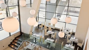 Un restaurante moderno con mesas y sillas de vidrio, ventanas amplias y lámparas esféricas colgantes.