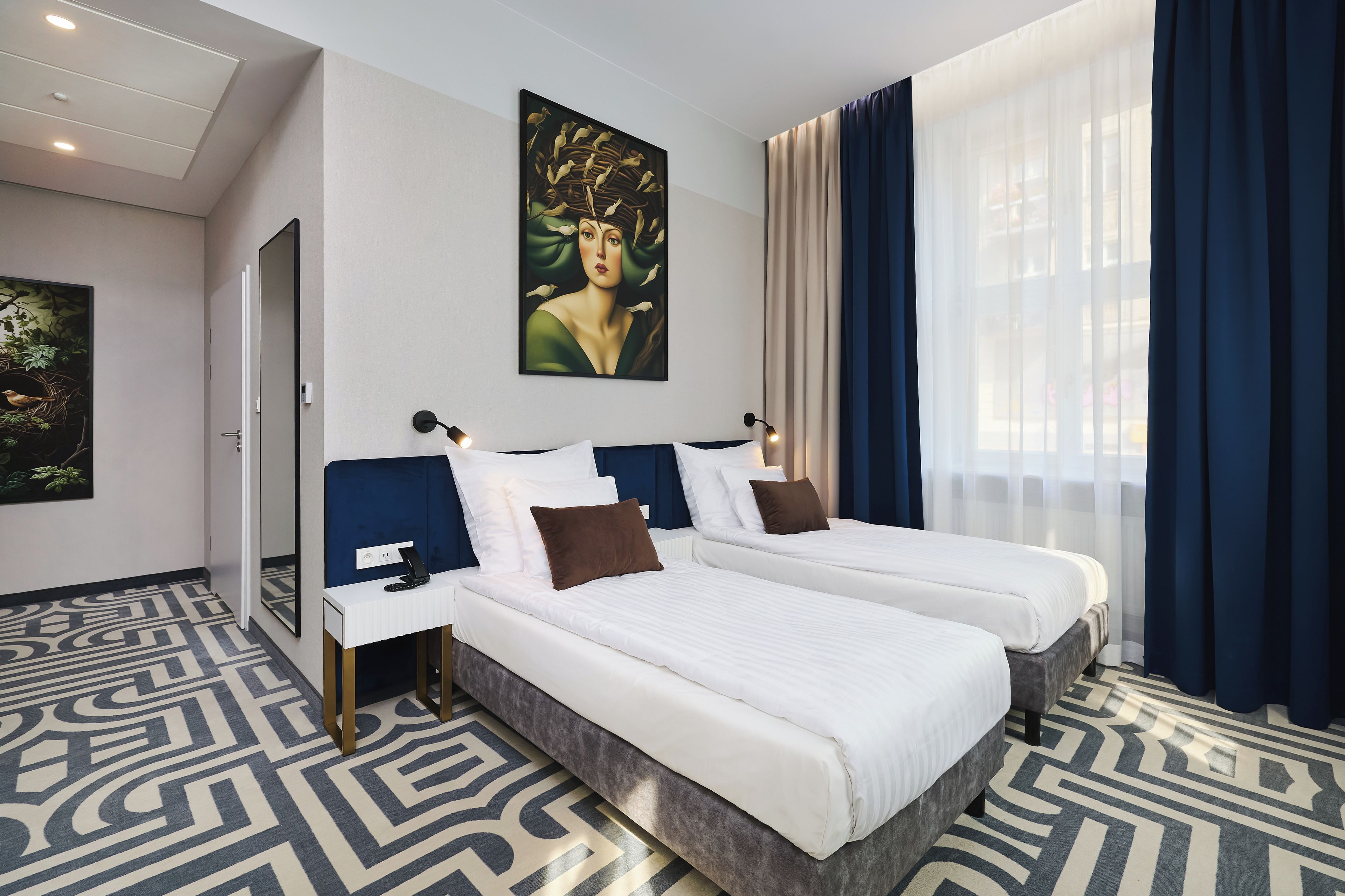 A hotel room with two beds, a large window with curtains, a painting on the wall, and a patterned carpet.