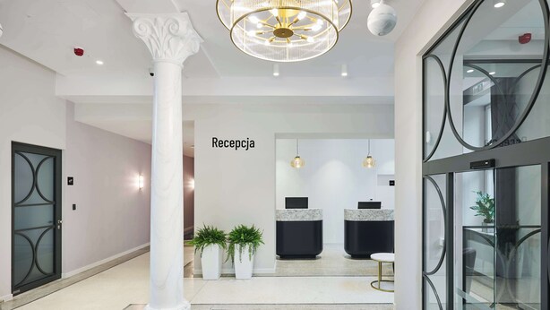 Uma área de recepção com balcão de mármore, plantas em vasos e uma escadaria que leva ao andar superior.