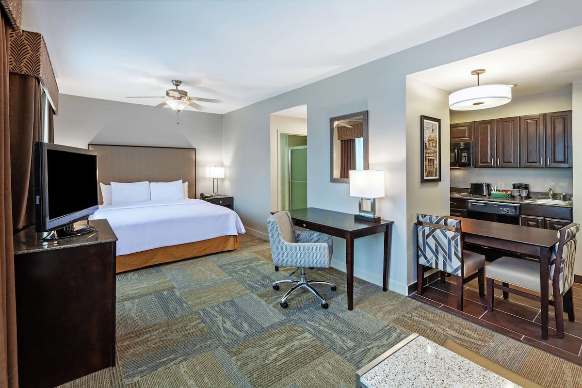 A hotel room with a bed, desk, chair, television, and kitchenette.