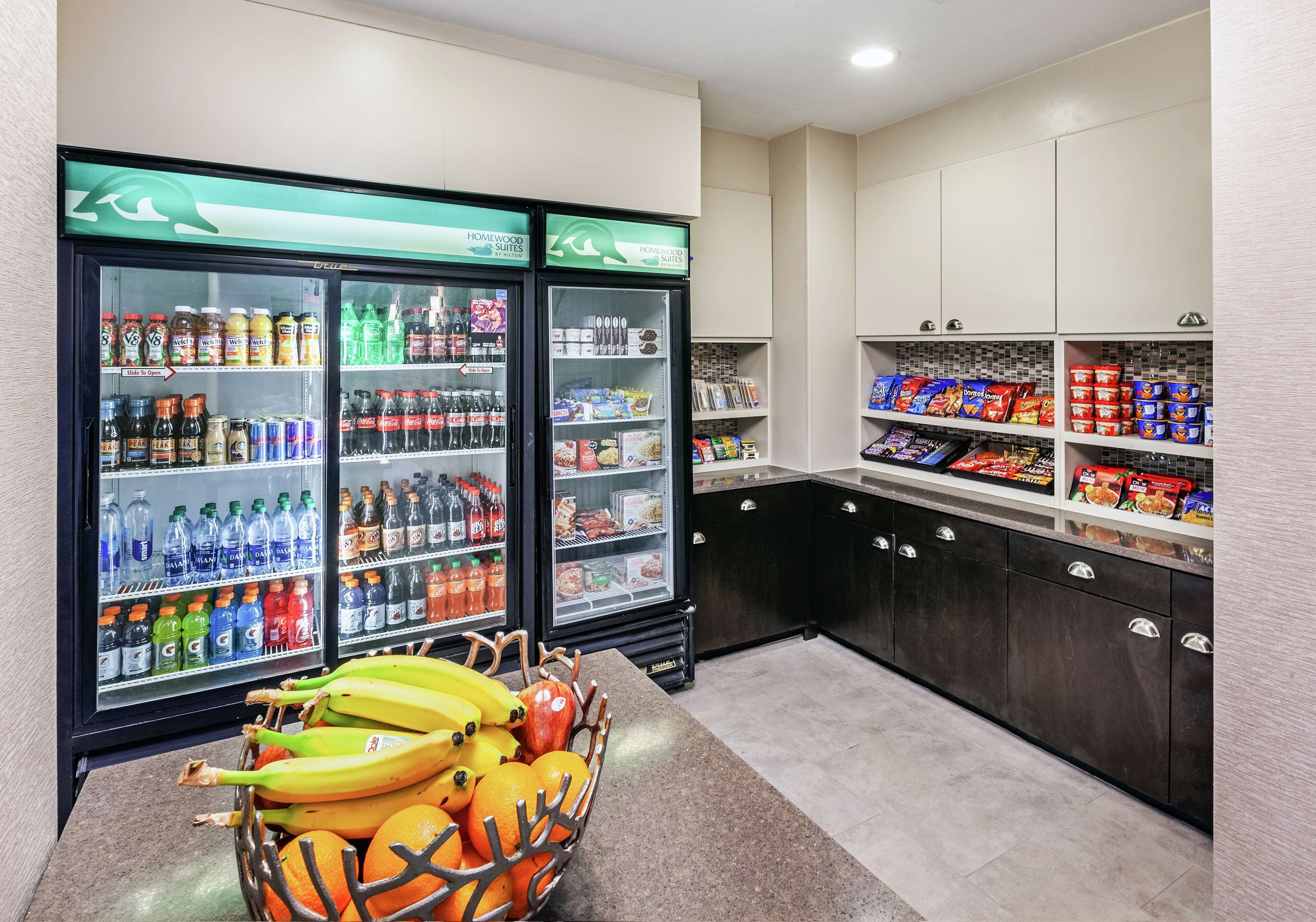 A kitchenette with a refrigerator stocked with beverages and snacks, a fruit basket on the counter, and cabinets with food items.