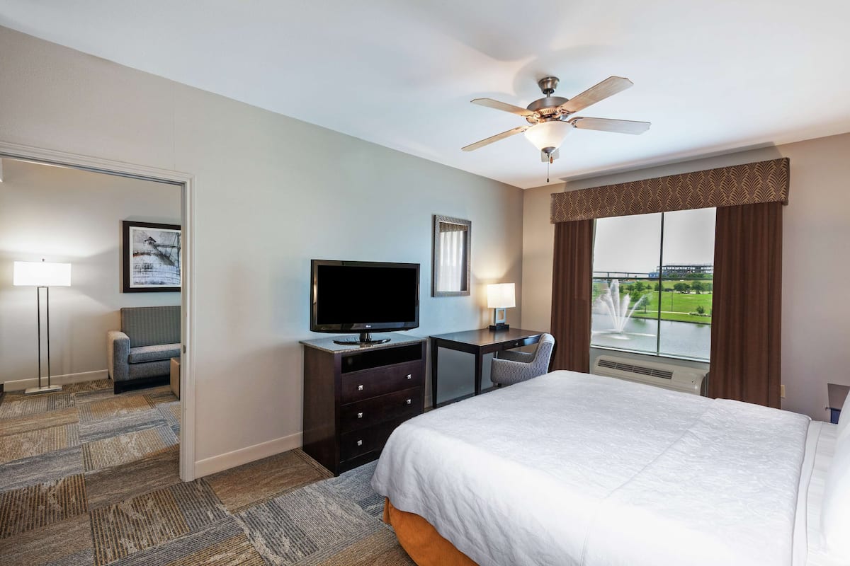 A hotel room with a bed, a desk, a television, a chair, a ceiling fan, and a large window with a view of a body of water.