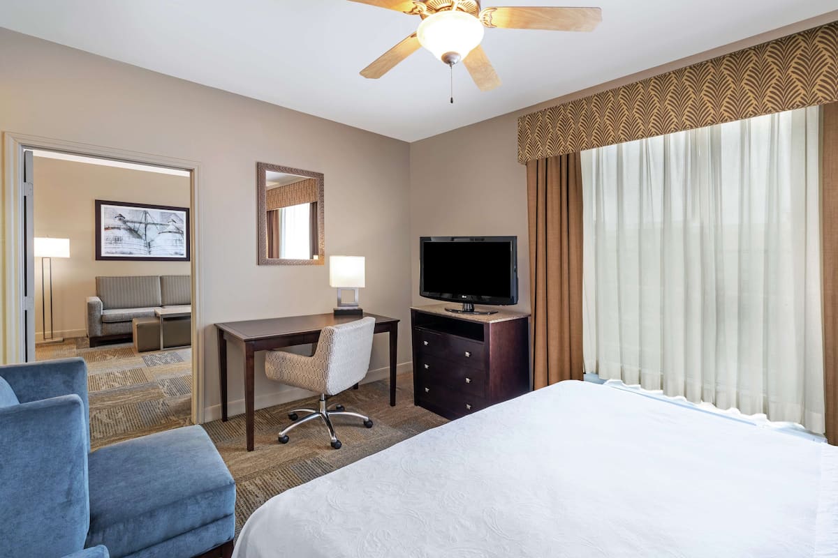 A hotel room with a bed, a desk with a chair, a television, a ceiling fan, and a large window with curtains.