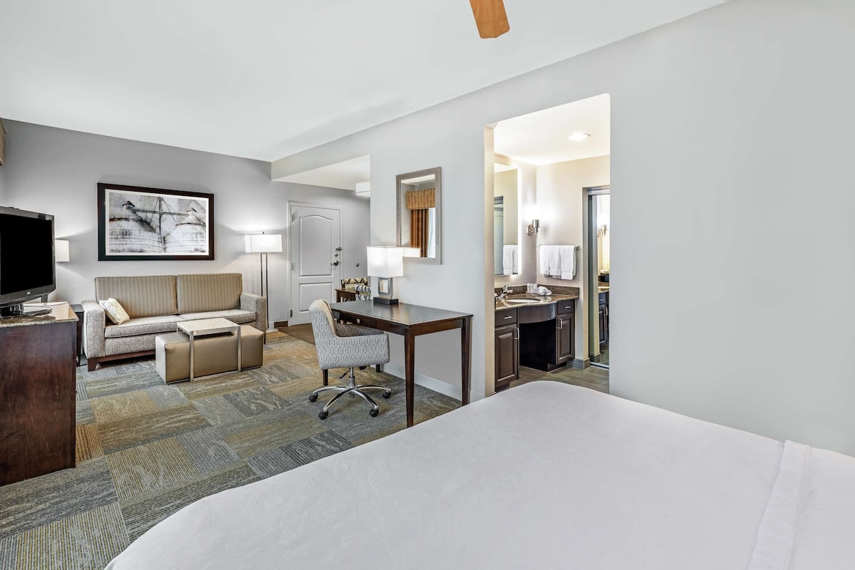 A hotel room with a bed, a desk, a sofa, a chair, a television, a lamp, and a bathroom visible through an open door.