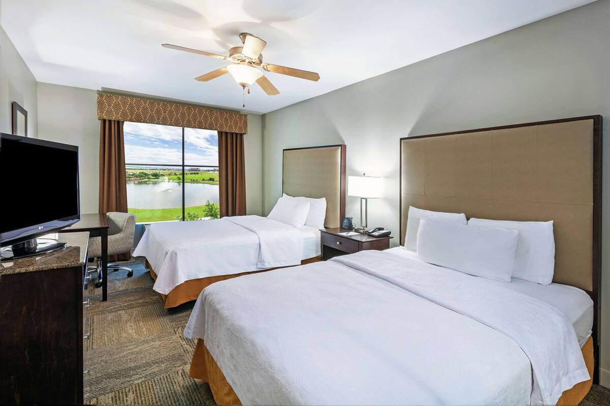 Ein Hotelzimmer mit zwei Betten, einem Fernseher, einem Schreibtisch, einem Stuhl, einer Lampe und einem Deckenventilator.